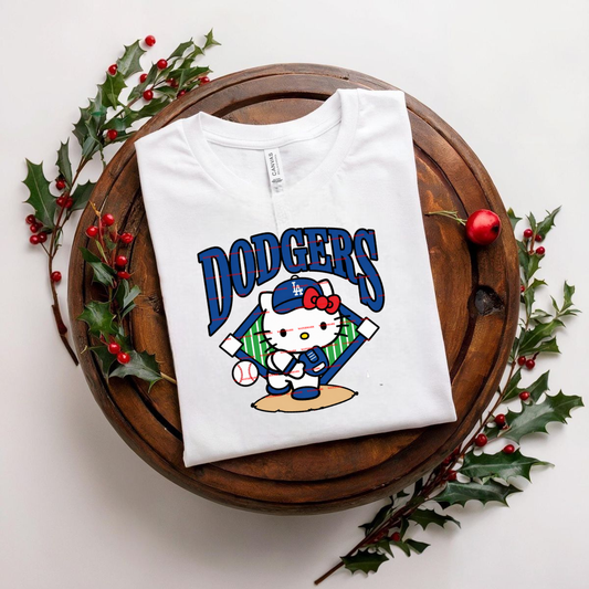 White t-shirt with 'Dodgers' design featuring Hello Kitty on a wooden surface with holly leaves and berries.